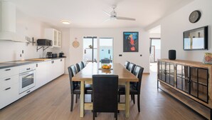 A modern kitchen with white cabinets, a wooden dining table, and black chairs. There is a ceiling fan, a clock on the wall, and a kitchen island with a microwave and toaster.