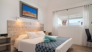 A neatly made bed with a geometric patterned bedspread, a wooden headboard, a nightstand, a wall-mounted painting, and a window with curtains.