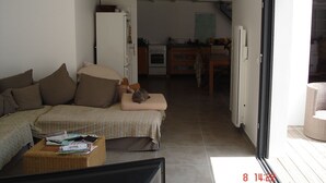 Un espacio habitable compacto con un sofá, una mesita con revistas, un televisor y un ventanal corredero que da a una cocina y comedor.