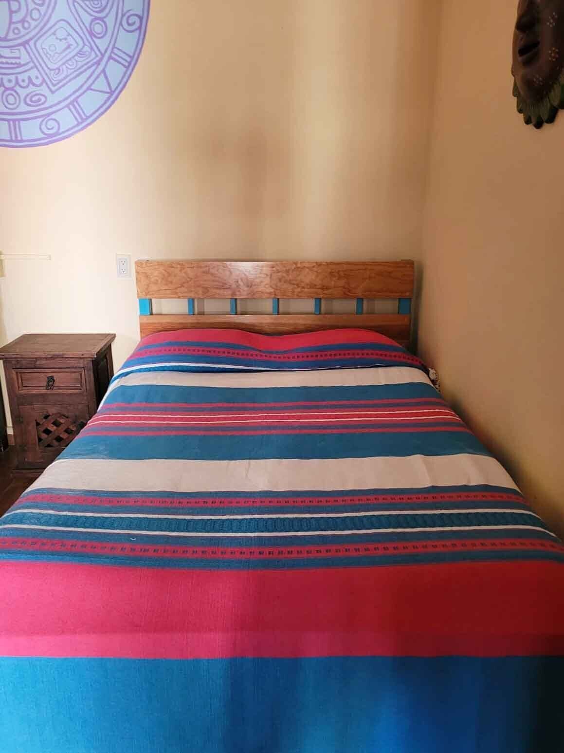 Una cama con colcha a rayas, cabecera de madera y mesita de noche de madera con cajón.