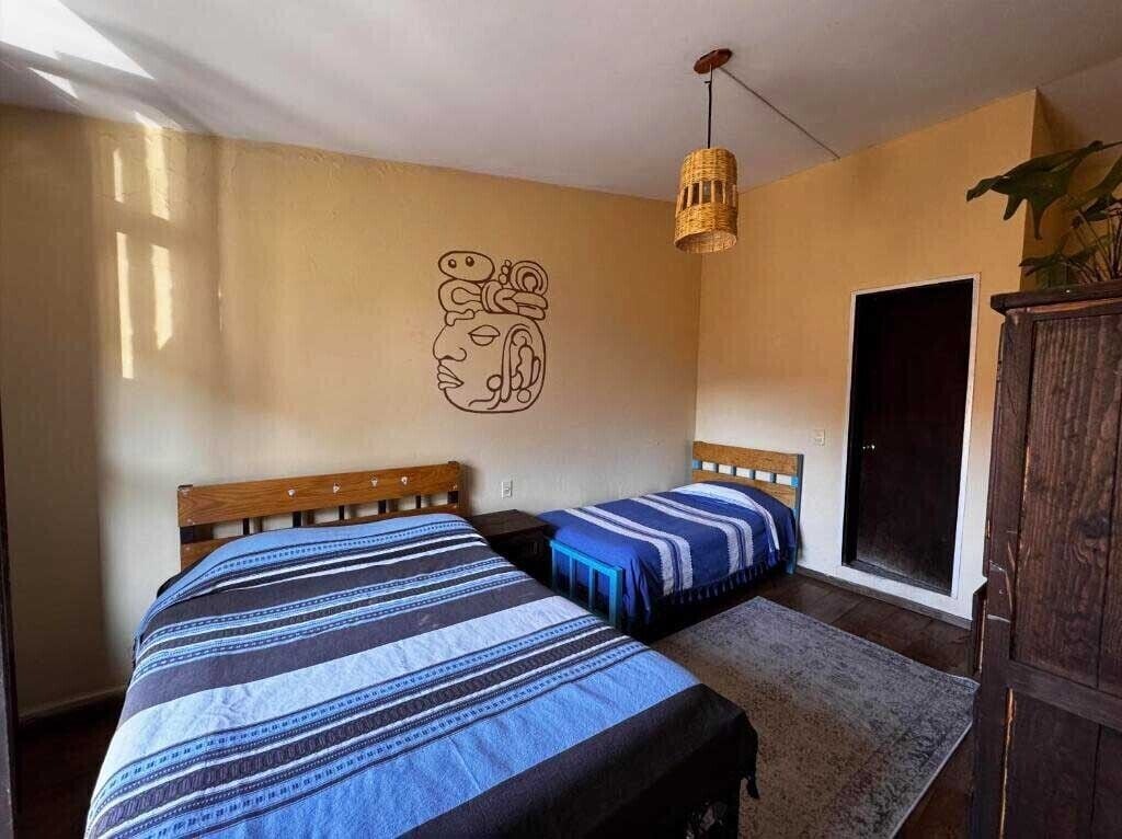 A room with two beds, a wooden headboard, a woven light fixture, a door, and a wall art piece.