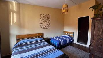 A room with two beds, a wooden headboard, a woven light fixture, a door, and a wall art piece.