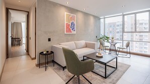 A modern living room with a sofa, a coffee table, and a dining area with chairs. There is a painting on the wall and a view of buildings through large windows. - Premium 1BR Apt with Jacuzzi & A/C in El Poblado (Medellín)