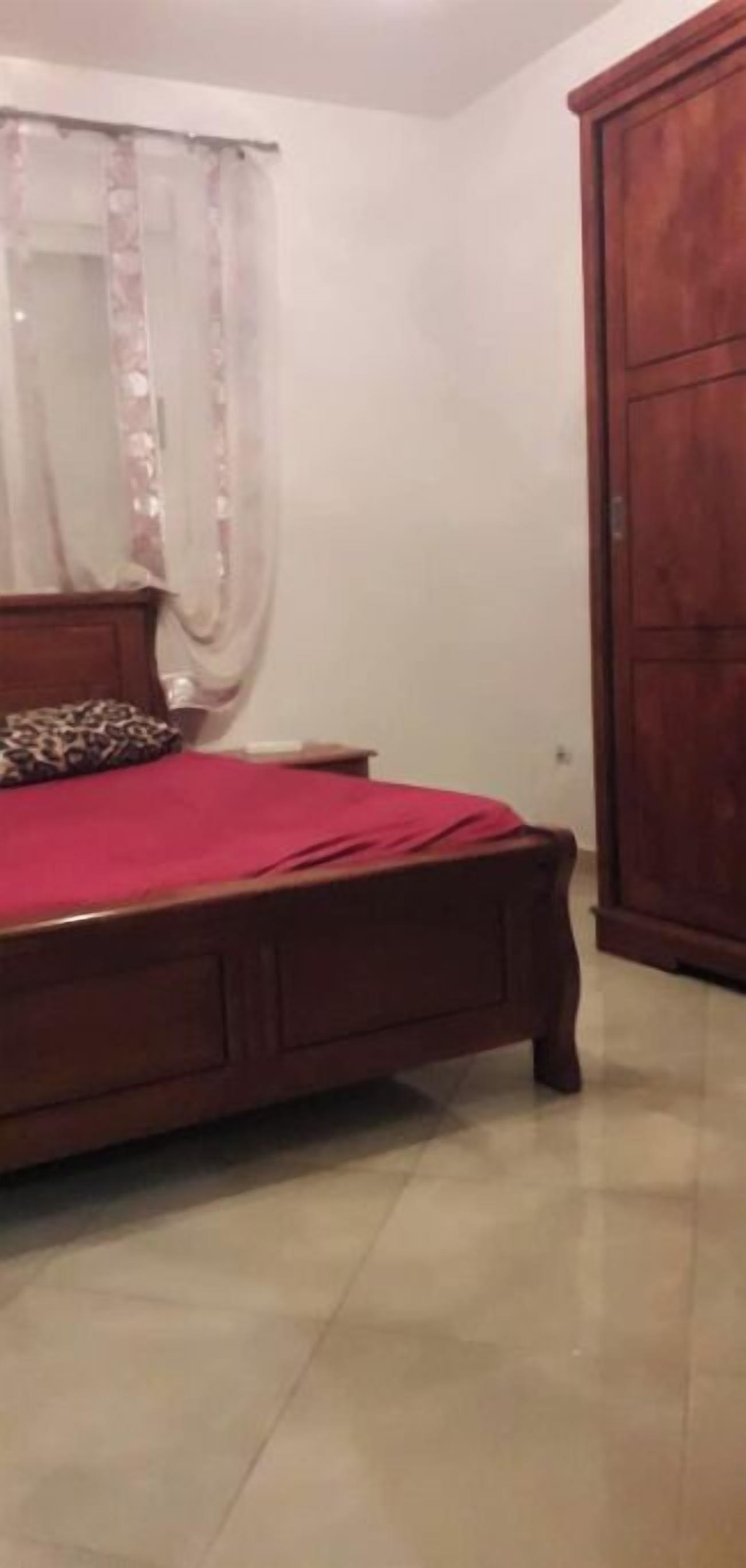 A bedroom with a wooden bed, a red bedspread, a wooden wardrobe, and a window with sheer curtains.