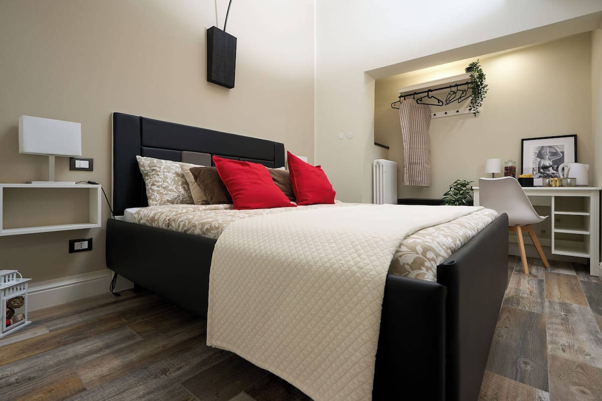 Un dormitorio moderno con cabecera negra, cama con colcha color beige, almohadas rojas y una mesita de noche con lámpara.