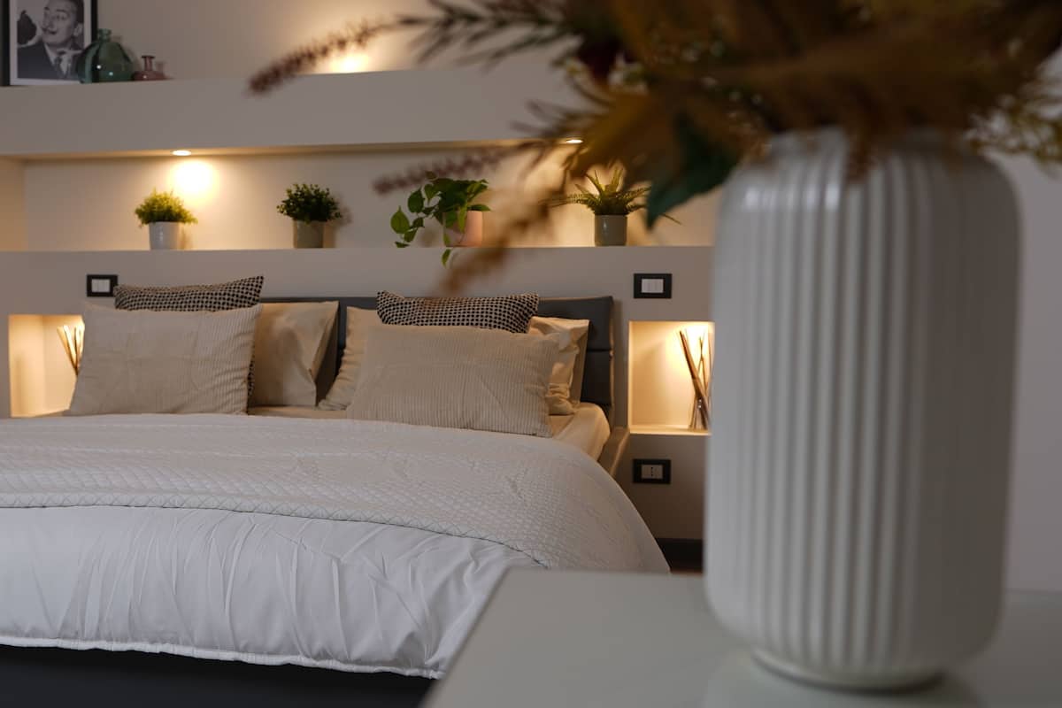 Una cama bien tendida con edredón blanco, una mesita de noche con un florero y un estante en la pared con objetos decorativos.