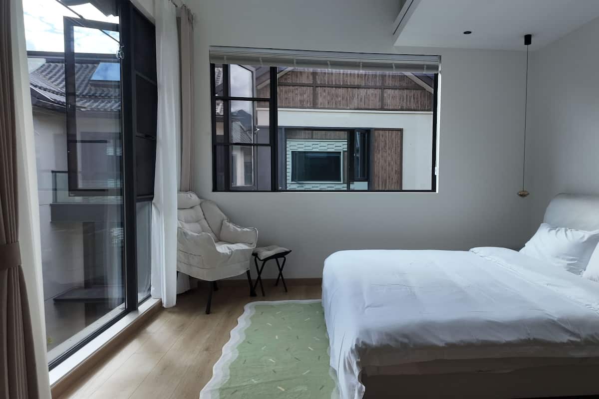 A bedroom with a bed, a chair, a window, and a view of buildings outside.