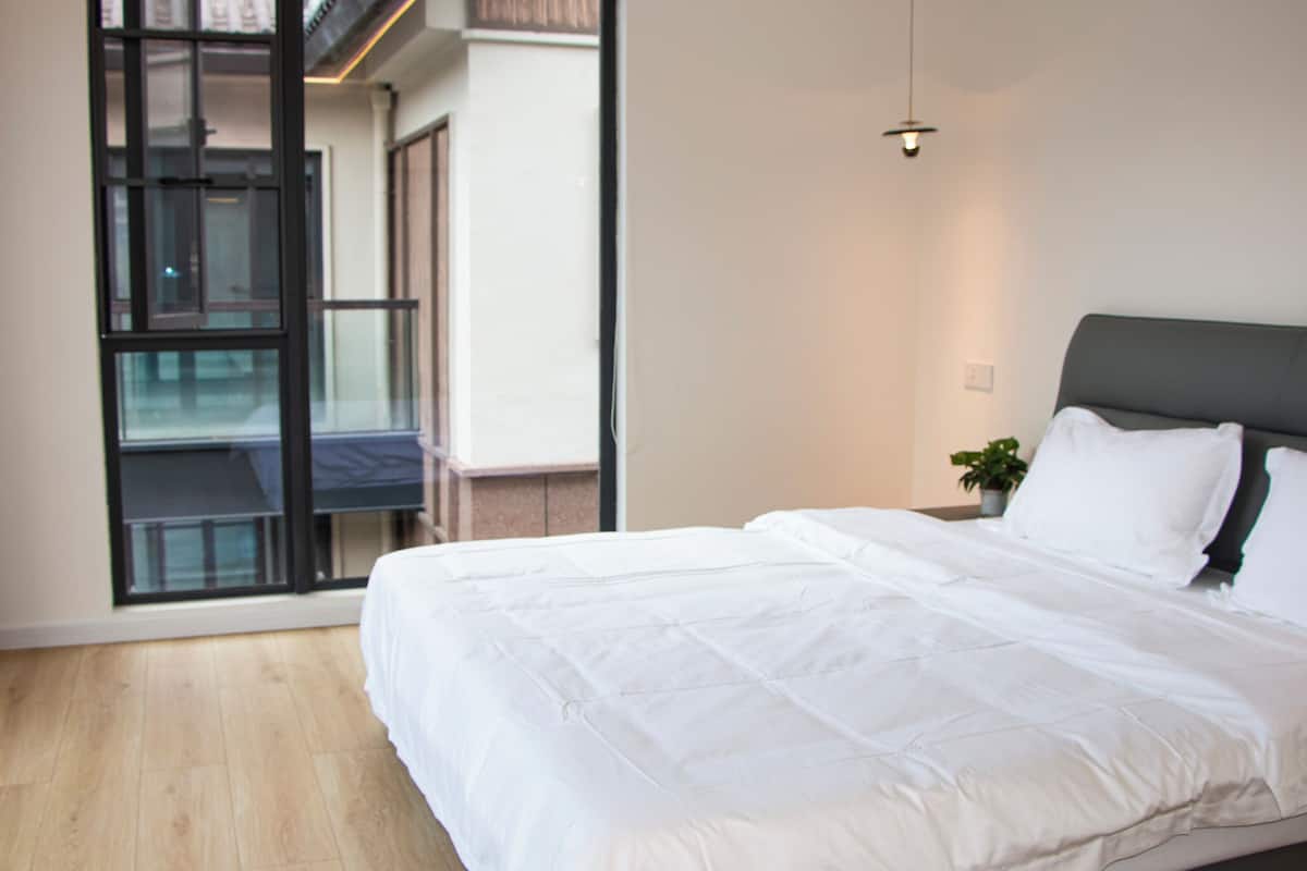 A modern hotel room with a large bed, a potted plant, and a view through a glass door to an outdoor balcony.