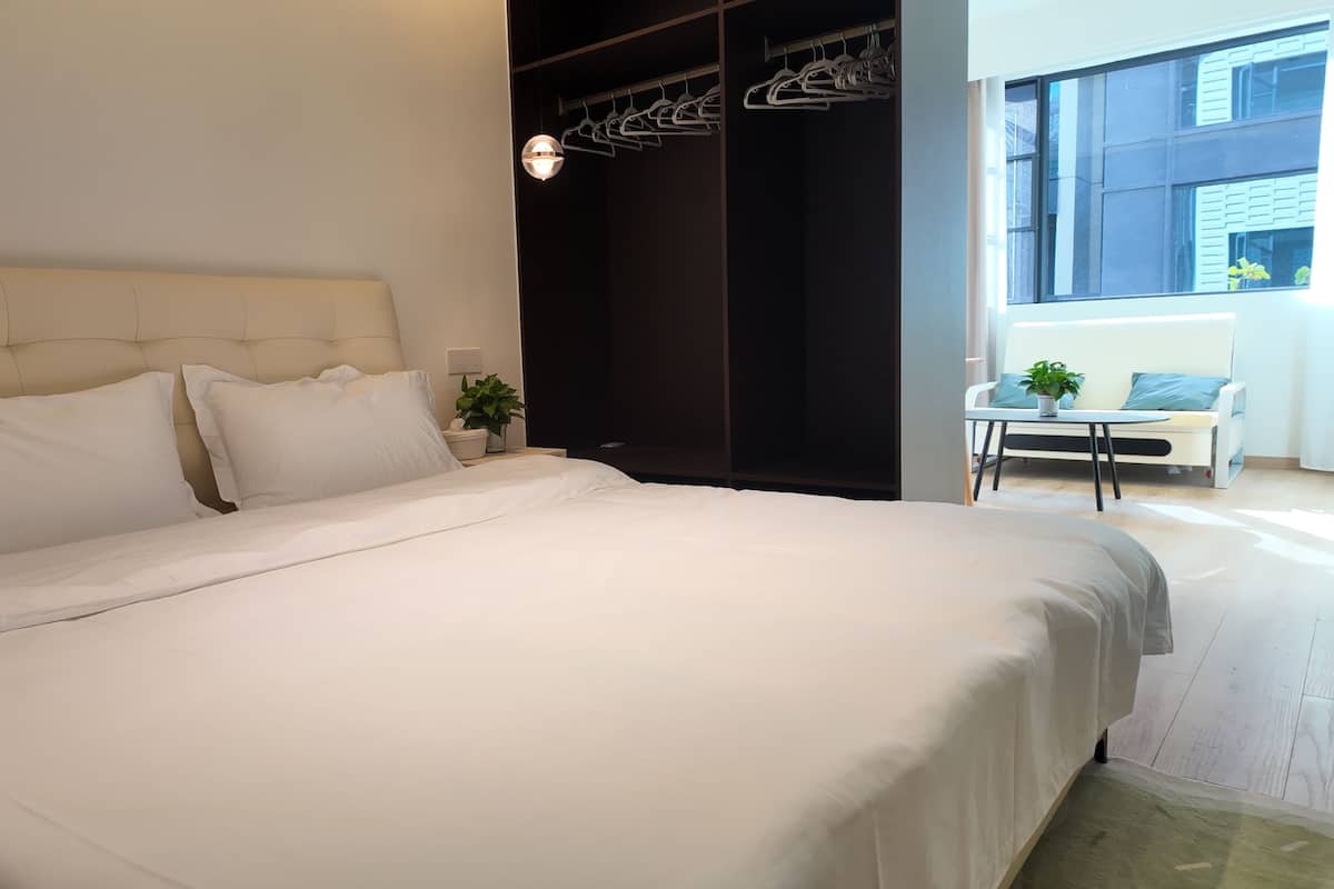A bedroom with a large bed, a bedside table with a plant, a hanging light fixture, and a view of the outside through a window.