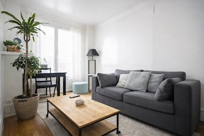 Olohuoneessa on harmaa sohva, puinen sohvapöytä, lamppu, ruukkukasvi sekä ruokailutila, jossa on pöytä ja tuolit.