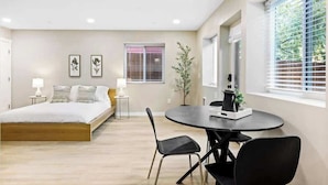 Un dormitorio moderno con cama, mesitas de noche, una mesa redonda de comedor con sillas, una ventana con persianas y una planta en maceta.