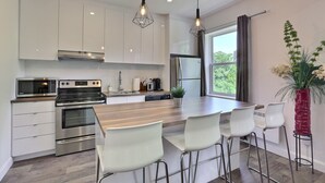 Una cucina moderna con mobili bianchi, elettrodomestici in acciaio inox, un'isola in legno con sgabelli da bar, illuminazione a sospensione e un'ampia finestra con tende.