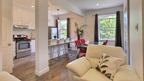 Una cucina moderna con mobili bianchi ed elettrodomestici in acciaio inossidabile, un'area pranzo con tavolo in vetro e sedie rosse, e un soggiorno con divano bianco e un cuscino decorativo.