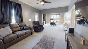 A living room with a sofa, armchair, and a ceiling fan. There is a dining area with a table and chairs in the background.