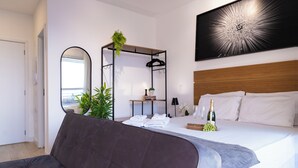 A modern hotel room with a large bed, a dark sofa, a wooden headboard, a wall-mounted mirror, a small table with a plant, a bottle of champagne, and a framed artwork. - Apt vista incrivel piscina e academia (São Paulo)