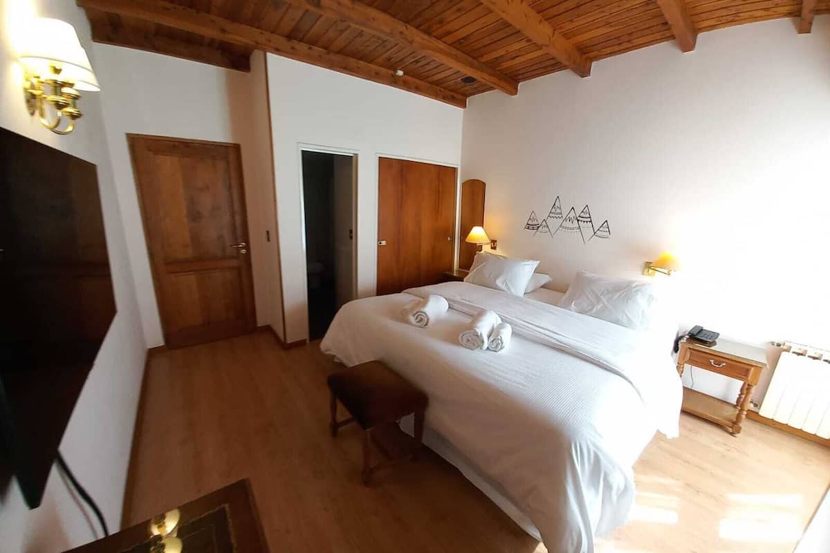 A hotel room with a wooden ceiling, a bed with white linens, a bedside table with a lamp, and a wall with a mountain design.