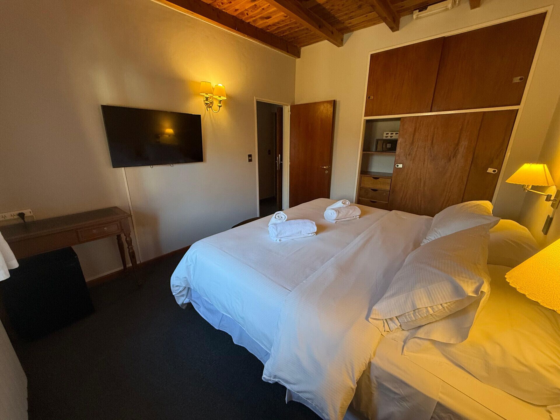A hotel room with a bed, a TV mounted on the wall, a wooden wardrobe, and a desk with a chair.