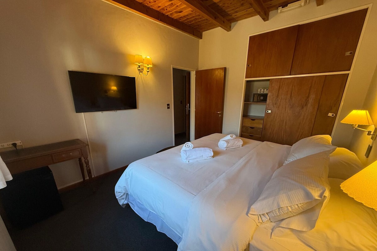 A hotel room with a bed, a TV mounted on the wall, a wooden wardrobe, and a desk with a chair.