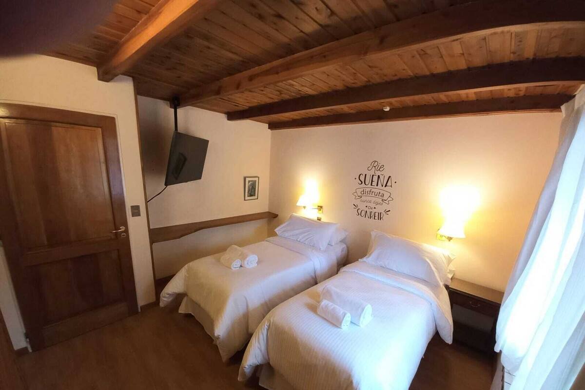A hotel room with two beds, wooden ceiling beams, a TV mounted on the wall, and a wall decoration with text.
