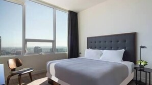 Ein Hotelzimmer mit einem großen Bett, einem Sessel, einem kleinen Tisch und Blick auf die Stadt durch große Fenster.