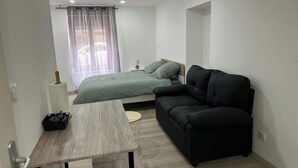 A modern, minimalist bedroom with a bed, a black sofa, a small table, and a window with curtains.