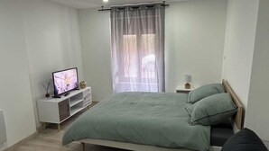 A modern bedroom with a bed, a TV on a white stand, a window with sheer curtains, and a bedside table with a lamp.