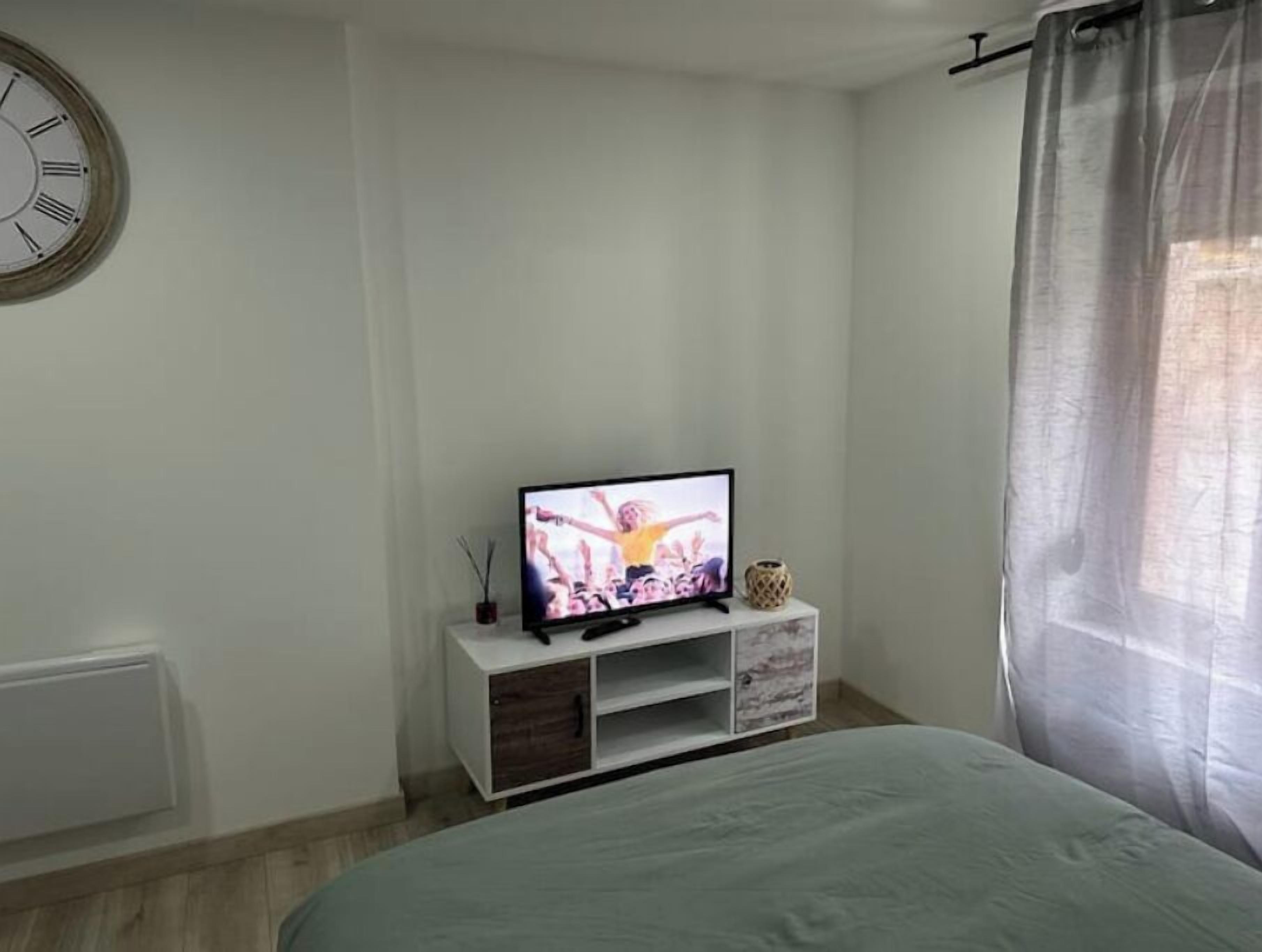 Une chambre à coucher avec un lit, un téléviseur posé sur un meuble blanc, une horloge au mur et une fenêtre avec des rideaux.