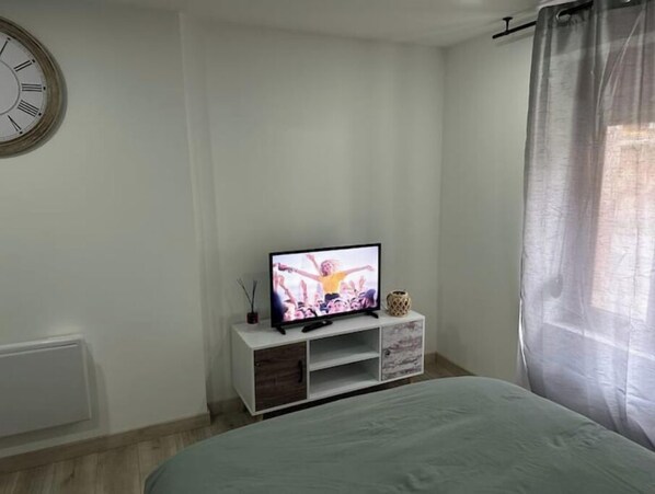 Une chambre à coucher avec un lit, un téléviseur posé sur un meuble blanc, une horloge au mur et une fenêtre avec des rideaux.