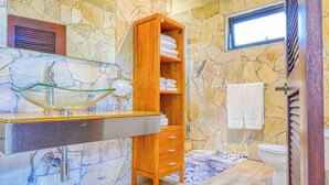 A bathroom with a stone wall, a wooden shelf with towels, a glass sink, and a toilet.