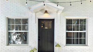 Uma casa de tijolos brancos com porta frontal escura, duas janelas com persianas e uma entrada com o tapete 'HELLO', além de luzes de Natal acima.
