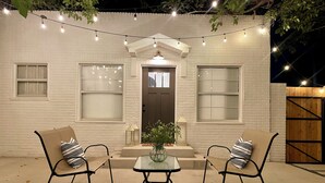 Uma casa de tijolos brancos com porta frontal escura, duas cadeiras com almofadas listradas, uma pequena mesa e luzes de cordão acima.
