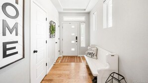 Un passadís amb una porta blanca amb la inscripció “HOME”, un banc amb un coixí i una planta a la paret.