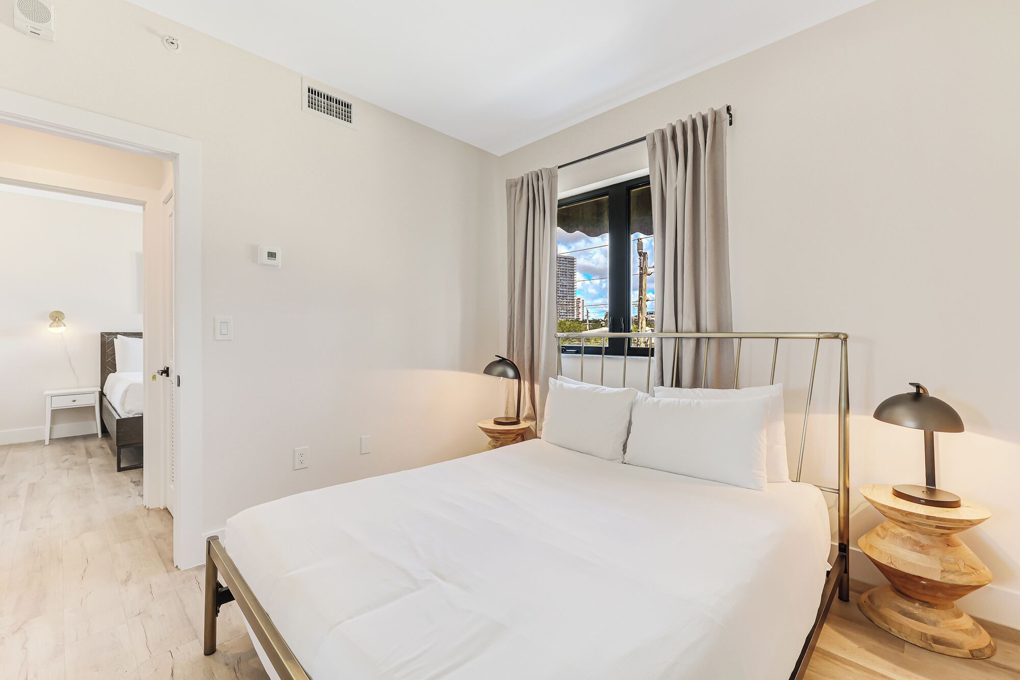 A modern bedroom with a large bed, bedside tables with lamps, a window with curtains, and a view of buildings outside.