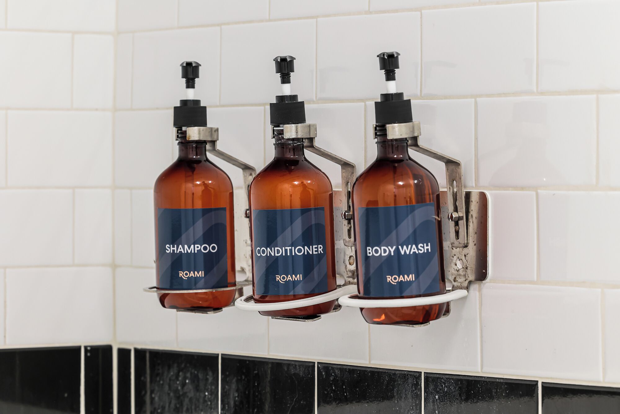 Three bottles of Roami products (shampoo, conditioner, body wash) mounted on a tiled wall.