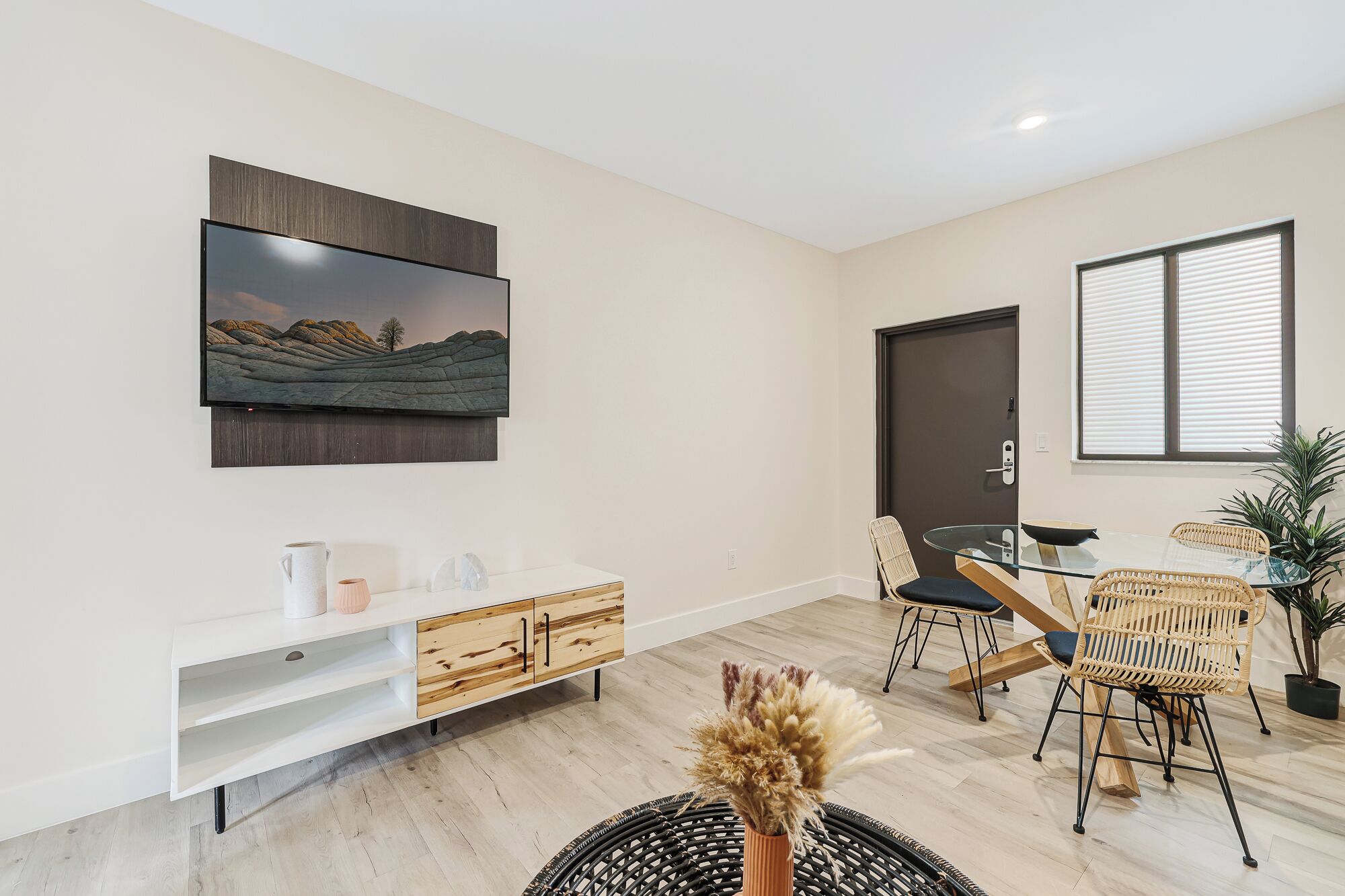 Una sala moderna con un televisor de pantalla plana montado en la pared, una mesa consola de madera, una mesa de comedor de vidrio con sillas de mimbre y una planta en maceta.