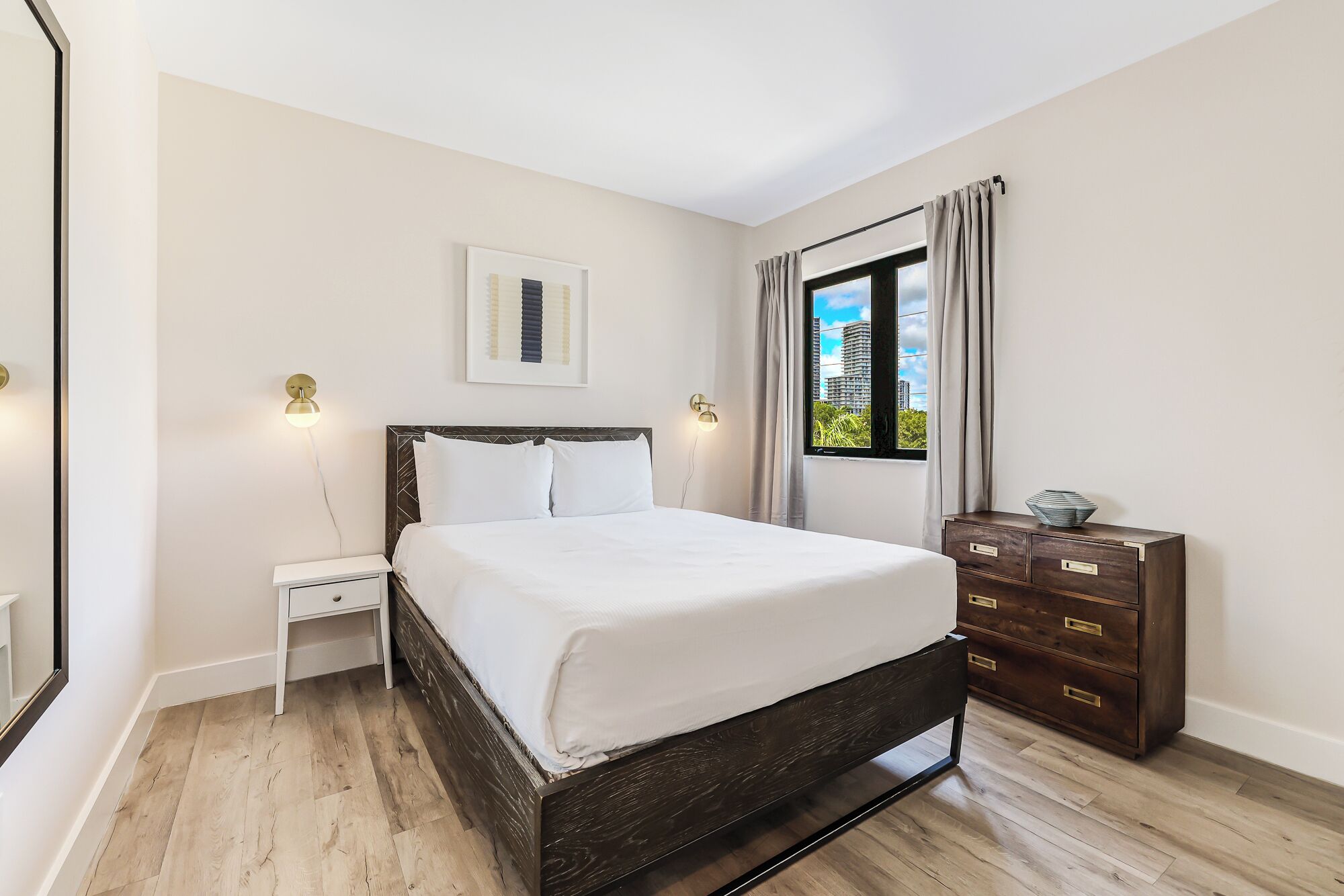 Un dormitorio con cama, mesitas de noche, cómoda, ventana con cortinas e imagen enmarcada en la pared.