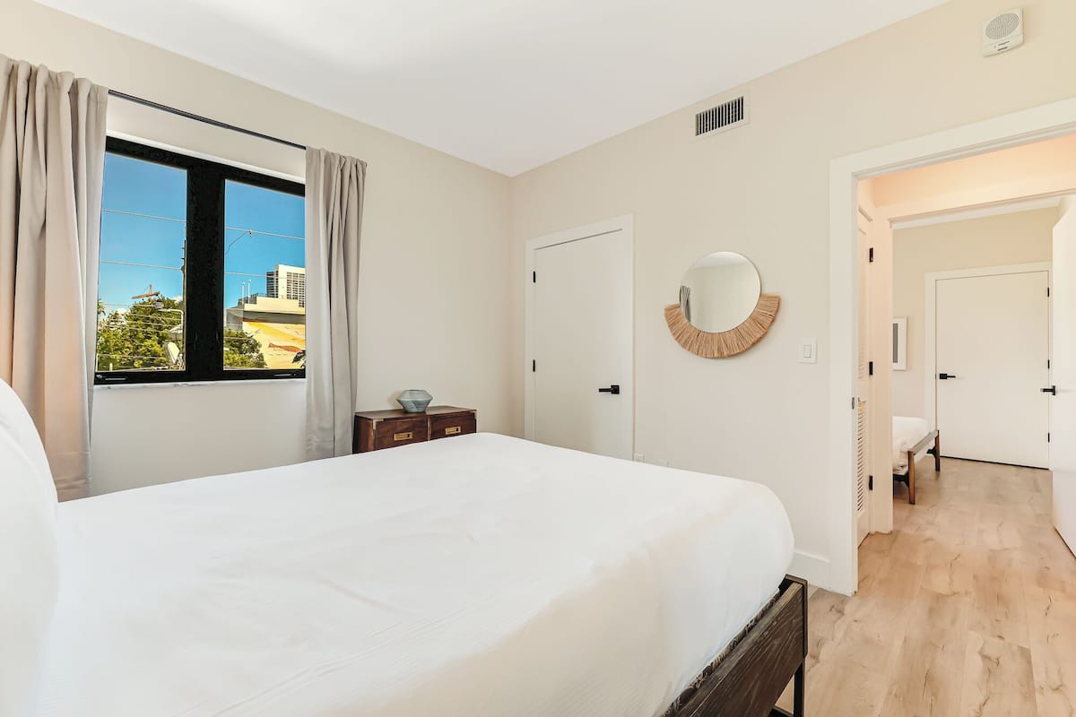 Un dormitorio con una cama grande, una ventana con cortinas, una mesita de noche, una puerta y un espejo en la pared.