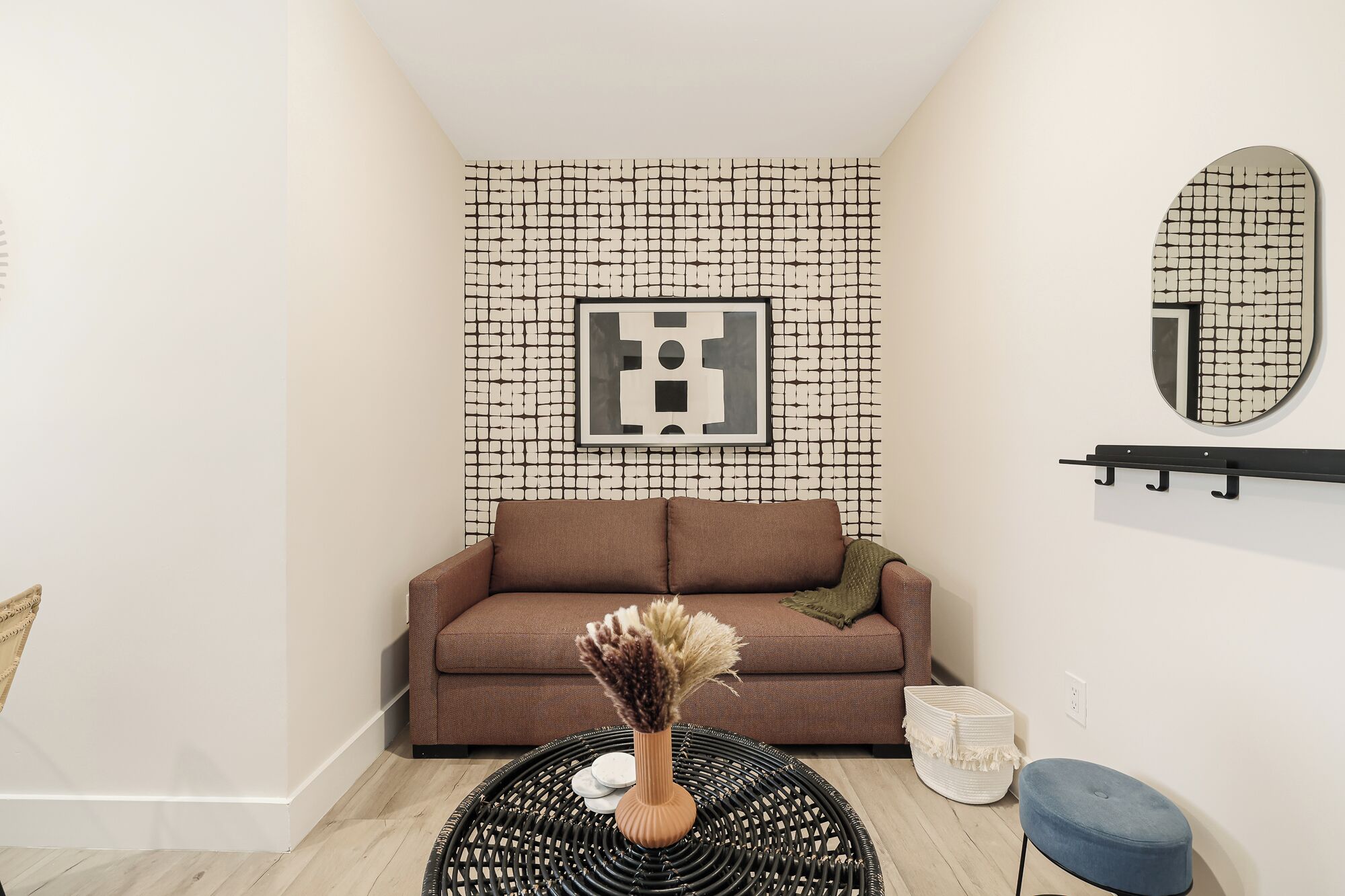 A modern living room with a patterned wall, a brown sofa, a round black wicker table, and a blue ottoman.