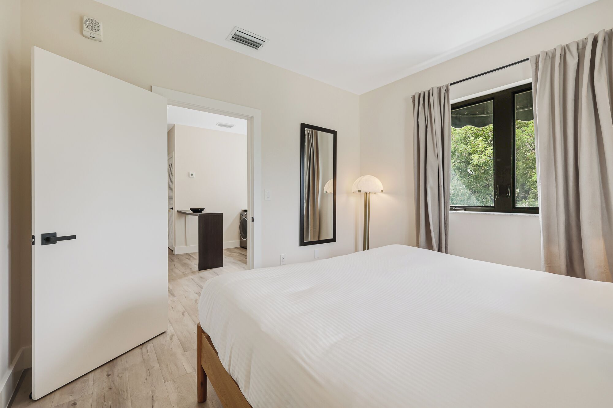 A hotel room with a bed, a bedside lamp, a window with curtains, a door, and a view of the outdoors.