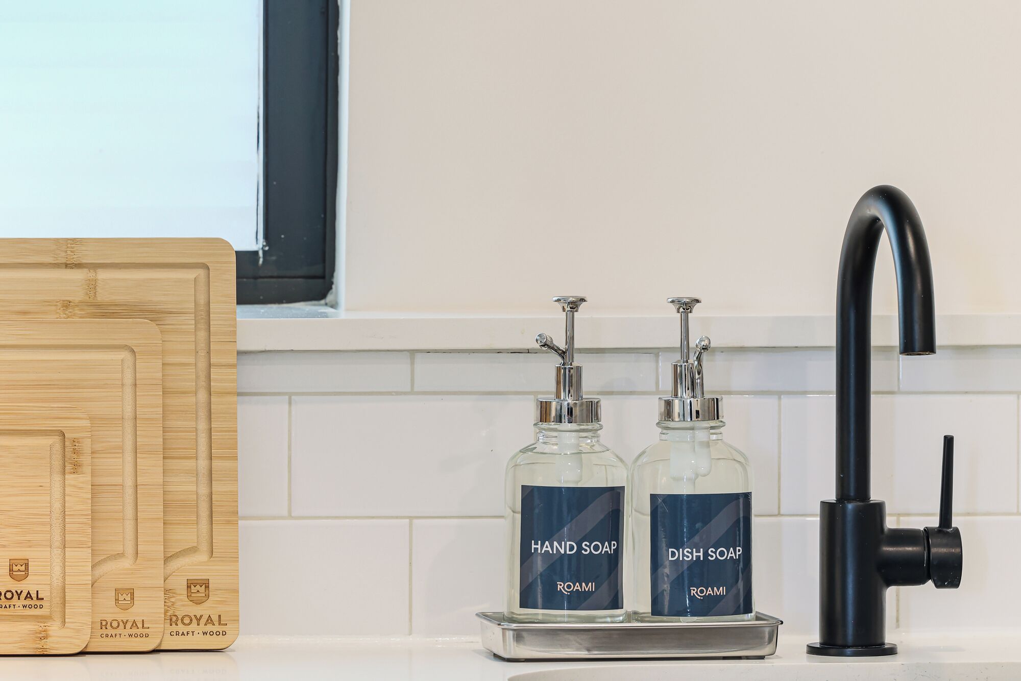 Uma cozinha com uma torneira preta, dois dispensadores de sabão rotulados 'Sabonete Líquido' e 'Detergente', e uma tábua de corte de madeira com a marca 'Royal Craft Wood'.