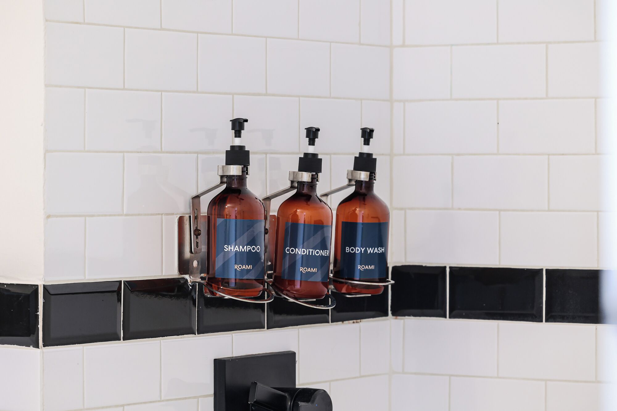 Three bottles of Roami products (shampoo, conditioner, body wash) mounted on a black and white tiled wall.