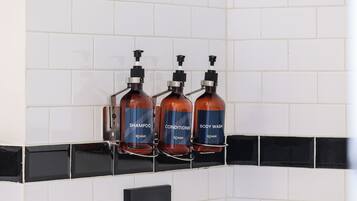 Three bottles of Roami products (shampoo, conditioner, body wash) mounted on a black and white tiled wall.