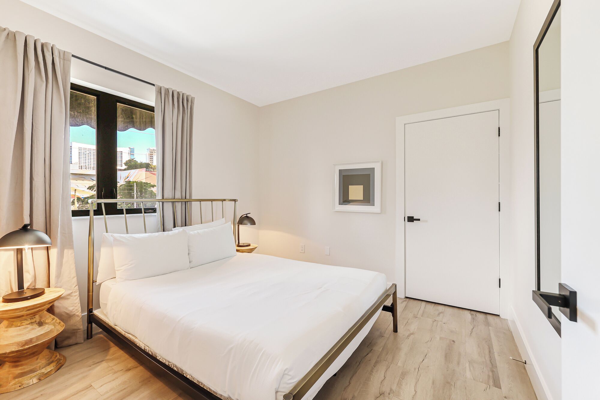 A modern hotel room with a bed, bedside table, lamp, and a view of the city through the window.