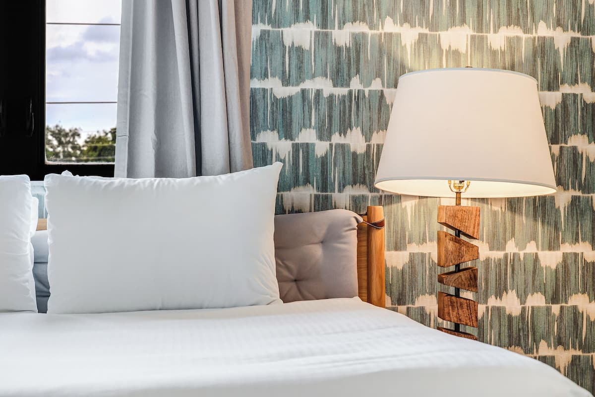 Una cama bien hecha con sábanas blancas, un cabecero tapizado en gris y una mesita de noche de madera con una lámpara. La pared presenta un papel pintado con líneas verticales y una ventana con cortinas.