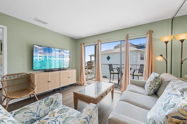 A modern living room with a flat-screen TV, wicker furniture, a sofa, and a glass-top coffee table. - Romar Beach 203: Beautiful Gulf View! (Orange Beach)