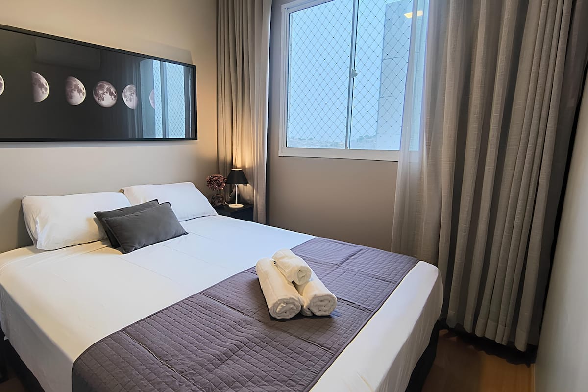 Quarto de hotel com uma cama grande, mesa de cabeceira com abajur, janela com cortinas e uma obra de arte na parede representando as fases da lua.