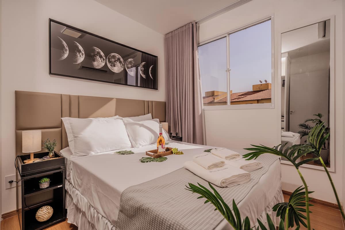 Cama bem arrumada com lençóis brancos, mesa de cabeceira com abajur, obra de arte na parede representando luas e janela com cortinas.
