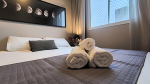 A hotel room with a bed, a wall-mounted artwork of the moon phases, a window with a view, and a neatly made bed with rolled towels. - Arena MRV, 2 quartos,vaga | Brazilian Corner (Belo Horizonte)