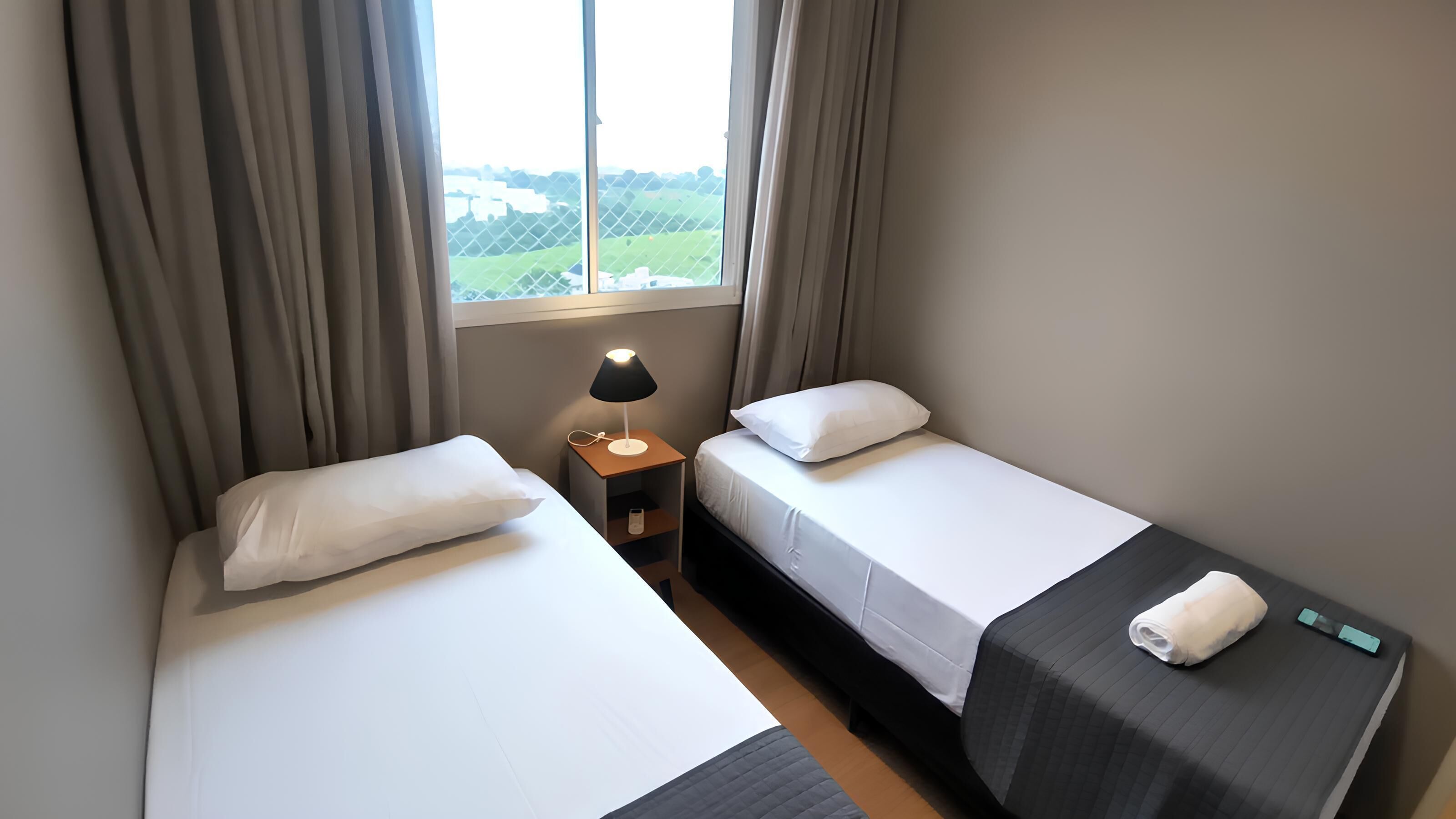 A hotel room with two single beds, a nightstand with a lamp, and a window with a view of the countryside.