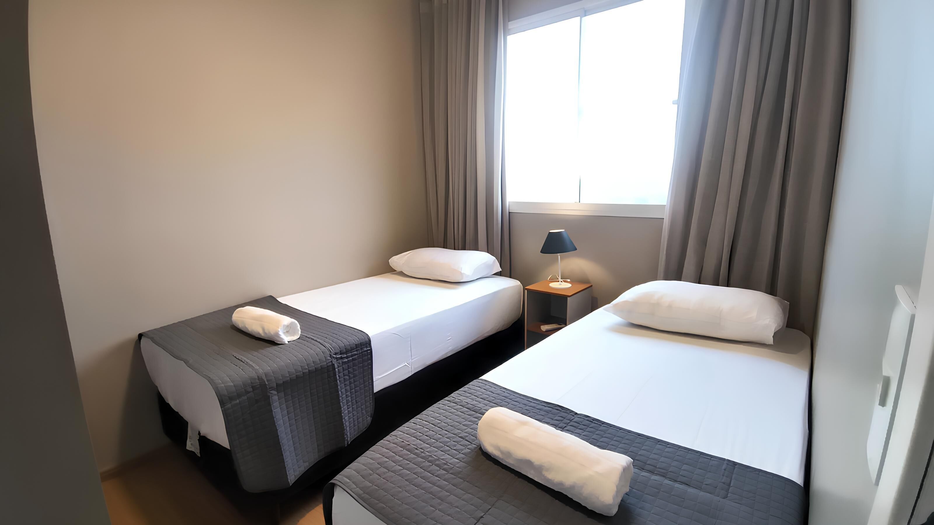 A twin room with two beds, each with a pillow and a rolled towel. There is a nightstand with a lamp between the beds.